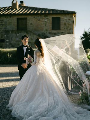 Bride in a ballgown with flowing veil and groom in tuxedo posing near a Tuscan stone villa.