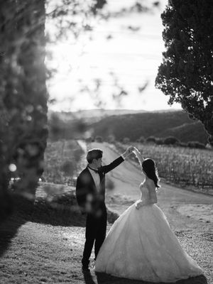 Black and white photo of groom spinning bride in ballgown on a Tuscan vineyard path.