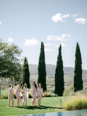Bridesmaids in blush pink outfits walking on green lawn with tall cypress trees in Tuscany.