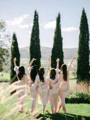 Bridesmaids in blush robes celebrating with raised arms before tall cypress trees in Tuscany.