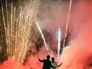 Bride and groom silhouetted against a spectacular fireworks display with colorful smoke at night.
