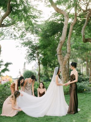 Bridesmaids in earth-tone gowns arrange bride's cathedral veil in lush garden wedding setting.