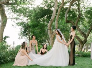 Bride in white gown with bridesmaids helping with train in lush garden setting.