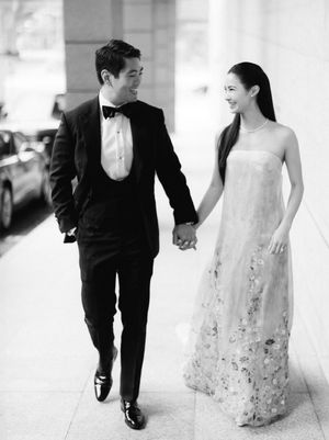 Black and white photo of couple holding hands, groom in tuxedo, bride in floral gown.