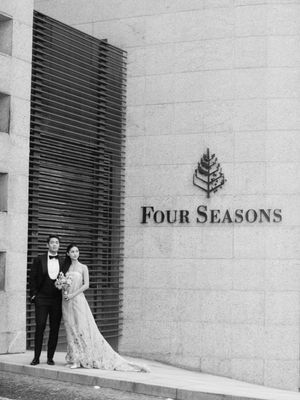 Bride and groom in formal attire posing outside Four Seasons Hotel in black and white photo.