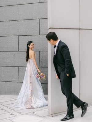 Bride in floral gown holds bouquet while groom in tuxedo peeks around a modern stone wall.