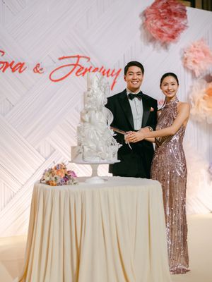 Smiling couple cuts elegant white wedding cake at reception with floral backdrop.