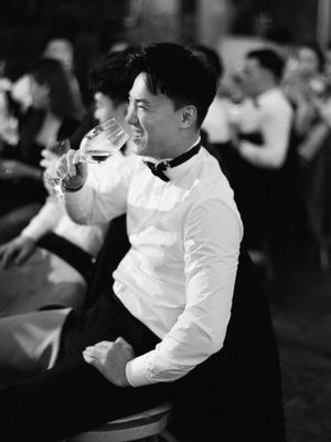 Joyful wedding guest laughs while drinking wine at reception in black and white photo.