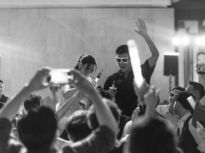 Black and white photo of a lively wedding reception with guests raising hands and phones around an energetic performer.