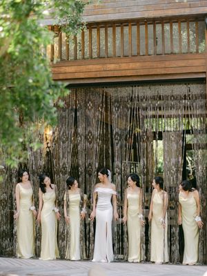 Bride in white gown stands with bridesmaids in gold dresses before a beaded curtain backdrop.