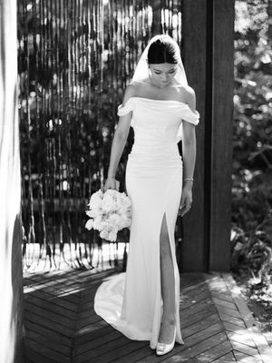 Elegant bride in off-shoulder white gown with slit holds bouquet in black and white portrait.