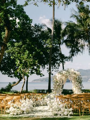 Stunning beachside wedding ceremony setup with white floral arch and wooden chairs on tropical lawn.