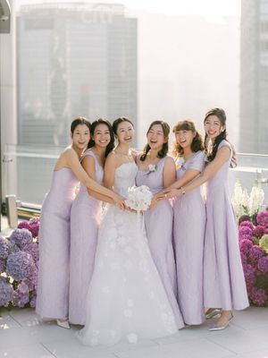 Bride in white gown holds bouquet surrounded by five bridesmaids in lavender dresses on a rooftop venue.