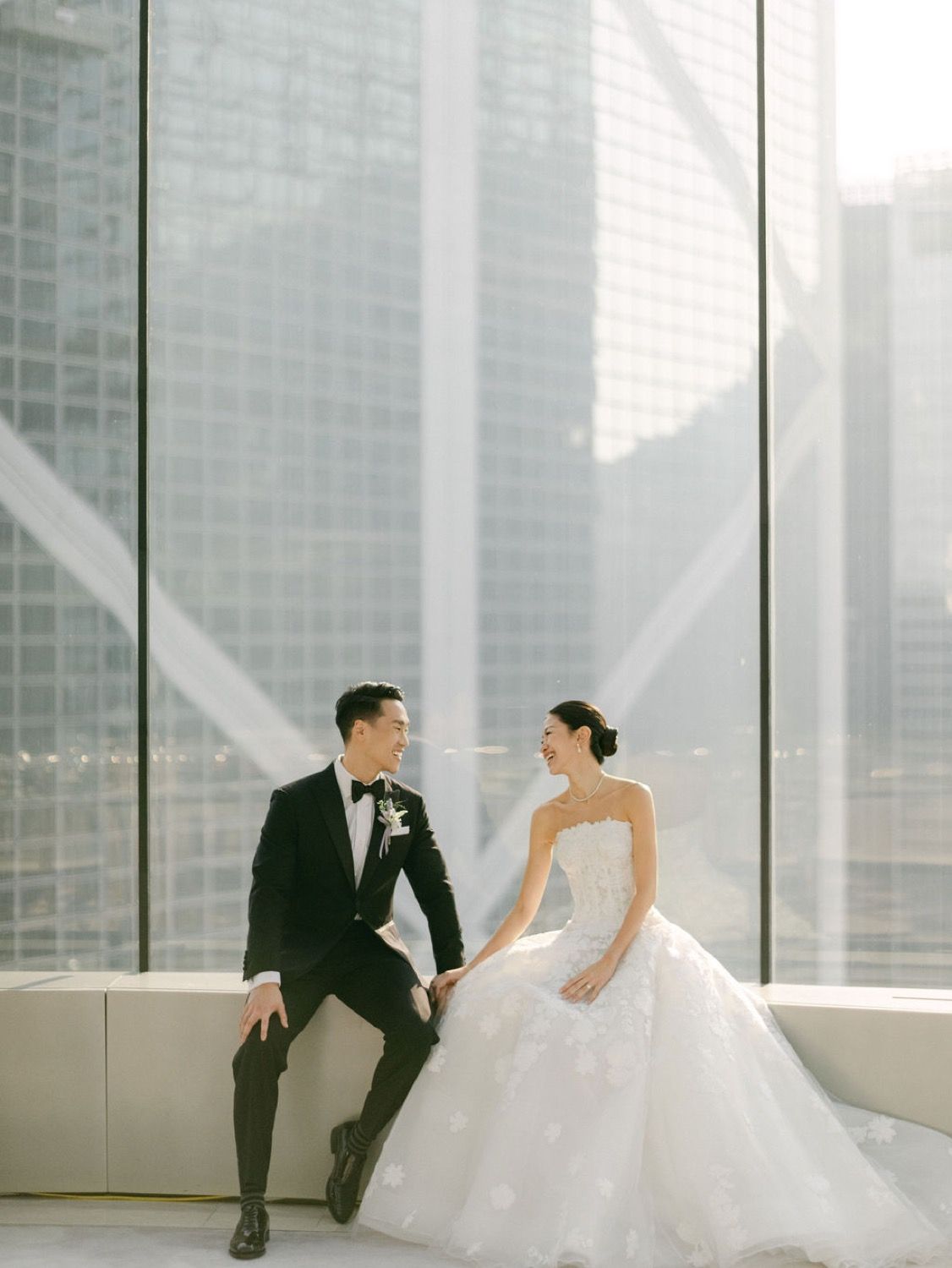 Elegant bride and groom share a romantic moment seated by floor-to-ceiling windows with city skyline backdrop.