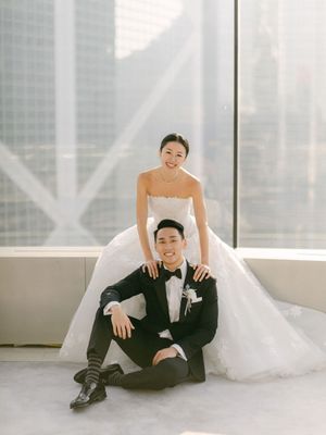 Bride in strapless ball gown poses with groom in tuxedo before stunning urban skyscraper window view.