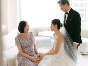 Bride in white gown shares tender moment with parents in formal attire at bright wedding venue.
