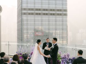 Couple exchanges vows at outdoor rooftop ceremony surrounded by purple floral arrangements and city skyline.