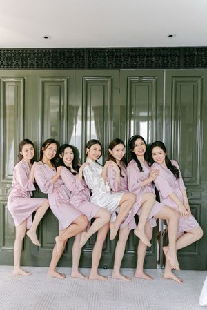 Seven bridesmaids in pink satin robes pose playfully against dark green cabinetry before wedding ceremony.