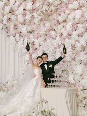 Joyful newlyweds celebrate with champagne bottles raised beside a champagne tower against a lush pink floral wall backdrop.