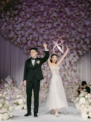 Celebrating couple raises hands before massive pink and white floral wall backdrop at wedding reception.
