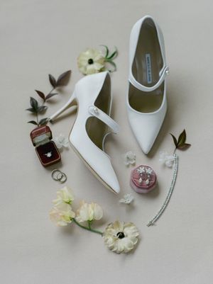 Elegant white pointed-toe bridal heels displayed with diamond rings, pearl earrings, and delicate white flowers on a neutral background.