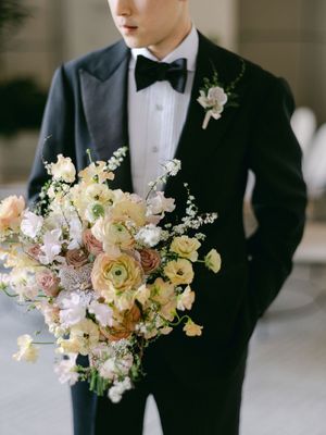 Dapper groom in black tuxedo holding a lush cascading bouquet of yellow, peach, and white ranunculus and wildflowers.