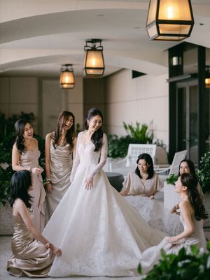 Bride in an elegant lace gown laughs joyfully as bridesmaids in champagne dresses help arrange her train on a covered terrace.