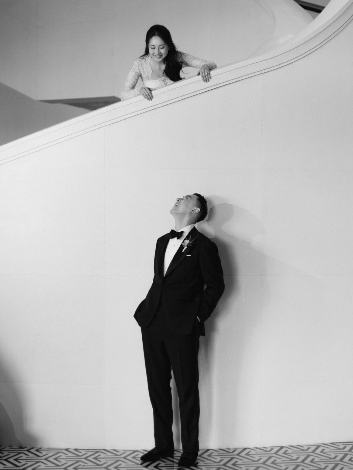Artistic black and white wedding photo of bride leaning over a curved staircase railing above her tuxedo-clad groom looking up.