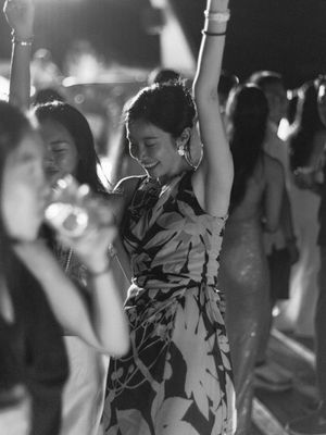 A woman in a floral dress dances joyfully with arm raised at an outdoor evening party in black and white.