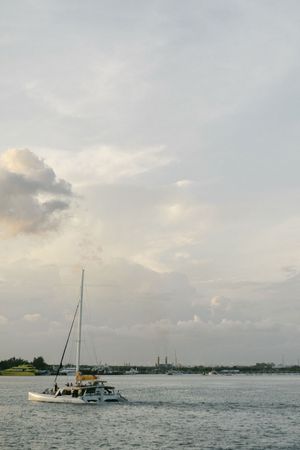 A white sailboat drifts peacefully on calm waters beneath a dramatic cloudy sky at golden hour near a distant shoreline.