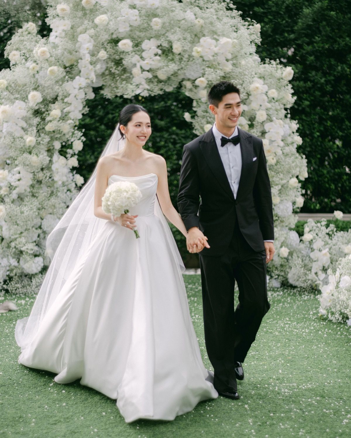 A smiling bride and groom walk hand-in-hand under a white floral arch at their outdoor wedding.