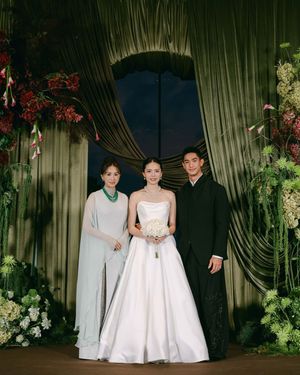 A bride in a white strapless gown poses with two guests before green floral wedding backdrop.