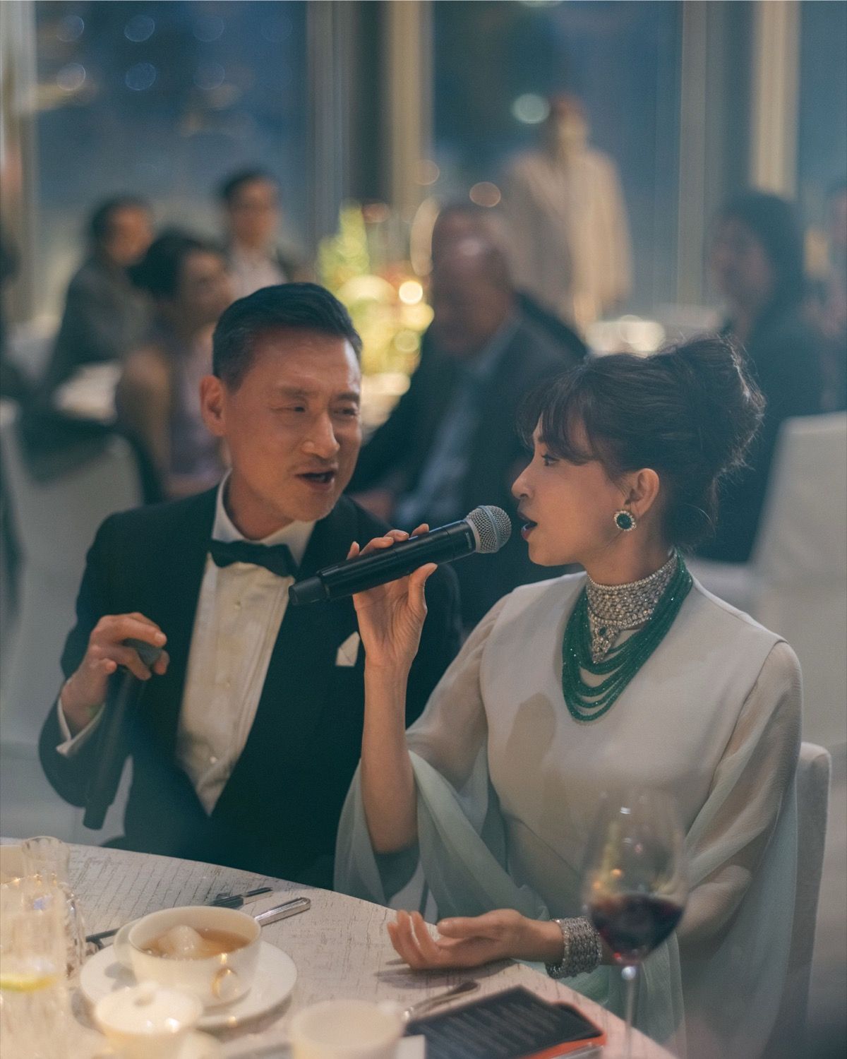 A man and woman share a microphone while seated at an elegant wedding reception dinner table.
