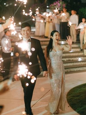 Newlyweds walk through sparkler exit, bride in sequin gown and groom in black tuxedo, smiling joyfully.