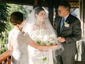A bride in a white veil receives her bouquet from parents during an outdoor wedding ceremony.