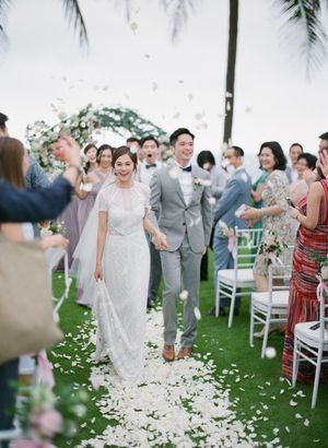 Newlyweds walk down outdoor aisle as guests toss white rose petals at tropical wedding celebration.
