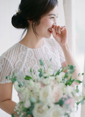 Joyful Asian bride in lace wedding dress laughs while holding white and pink floral bouquet near window.