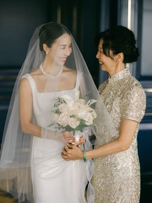 Bride in white gown and veil holding rose bouquet shares tender moment with mother in gold embroidered qipao.