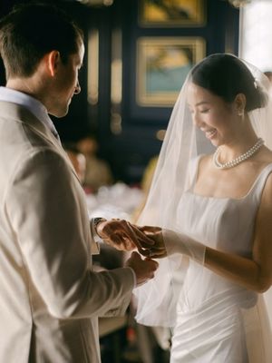 Bride in white gown and veil smiles as groom in beige suit places wedding ring on her finger at ceremony.