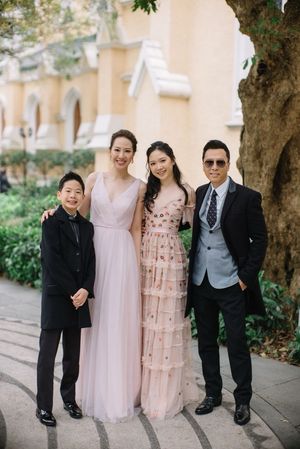 Family of four poses outdoors in formal wedding attire near a historic building with lush greenery.