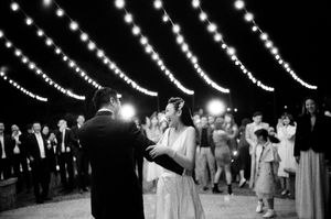 Bride and groom share first dance under string lights surrounded by guests in black and white photo.