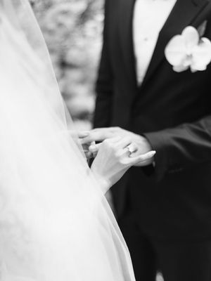 Black and white close-up of bride and groom holding hands, showing wedding ring and orchid boutonniere.