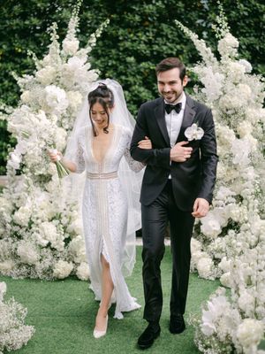 Bride and groom exiting ceremony under a stunning white floral arch with roses and baby's breath blooms.