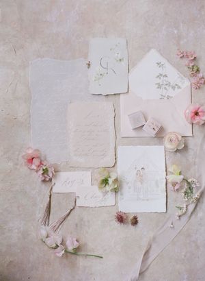 Romantic flatlay of handmade paper wedding stationery suite with pink florals, calligraphy, and velvet ring box.