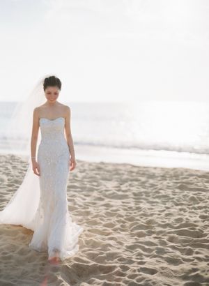 Elegant bride in strapless lace gown and veil walking on sandy beach.