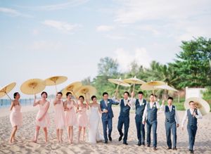 Wedding party on beach with parasols, bridesmaids in pink dresses and groomsmen in navy suits.