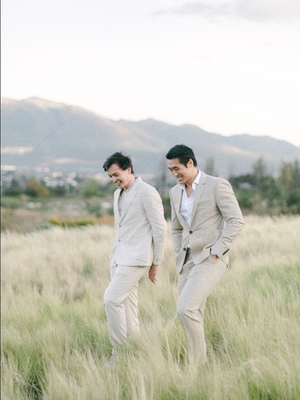 Two grooms in matching beige suits laughing together in a grassy field with mountain backdrop.