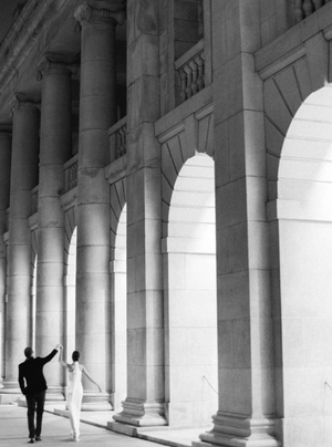 Bride and groom walk through grand classical stone colonnade in black and white photo.