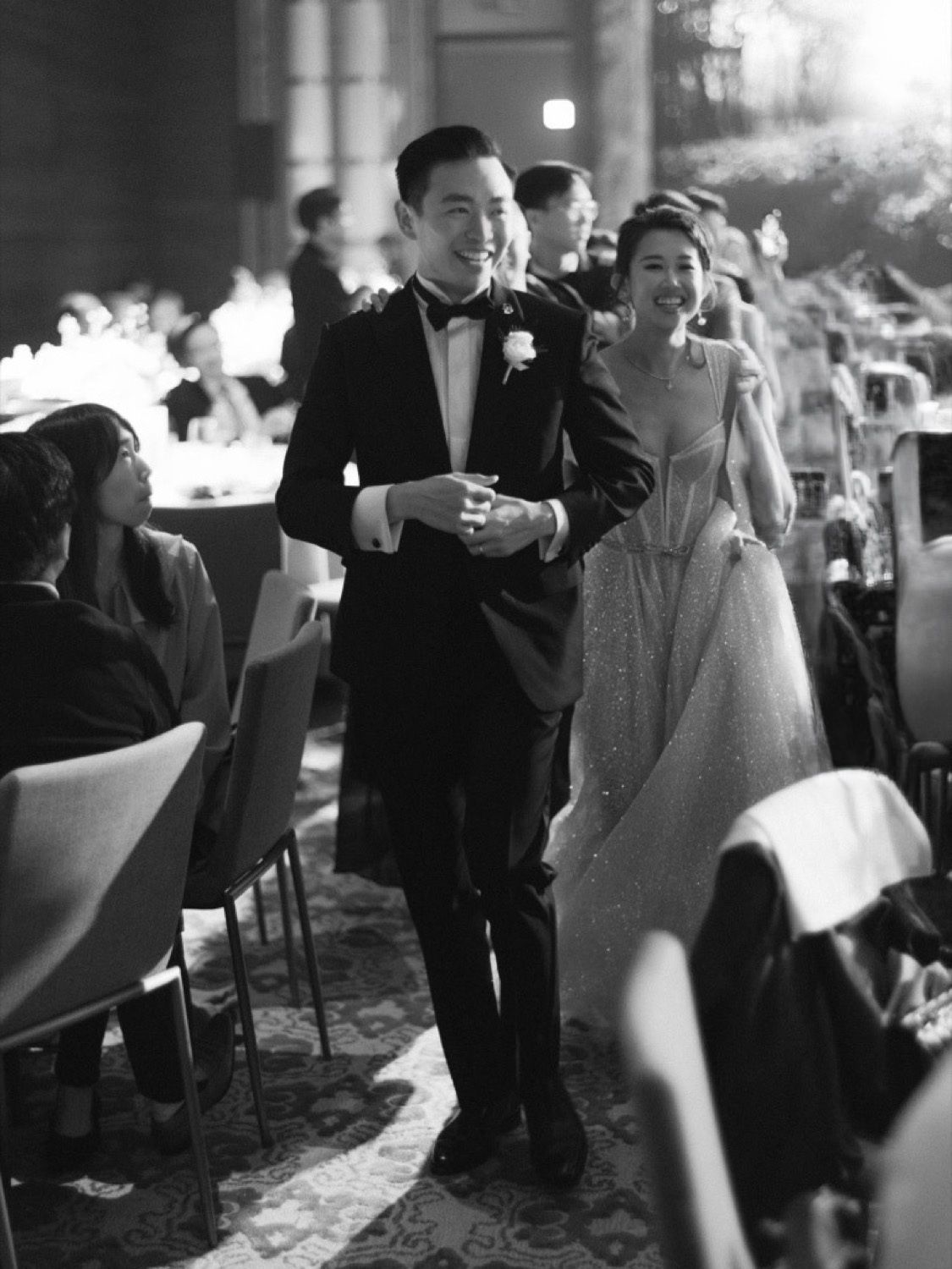 Black and white photo of a smiling couple in wedding attire walking through a reception hall.