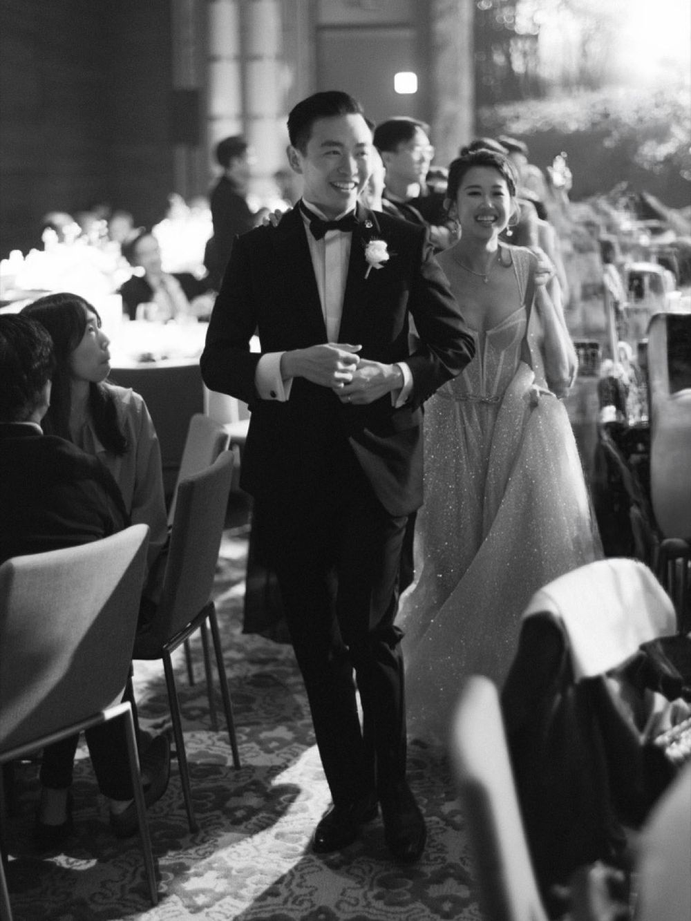 Black and white photo of a smiling couple in wedding attire walking through a reception hall.